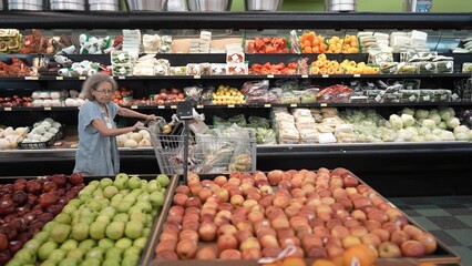 An elderly senior woman is grocery shopping in a vibrant store, carefully selecting fresh fruits and vegetables while pushing her cart.