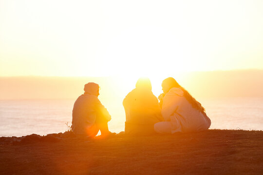 Three people sitting by the beach at sunset