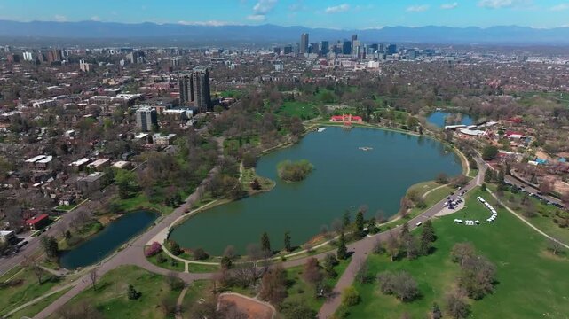 City Park Lodo Denver Colorado Lake Pavilion vibrant spring summer aerial drone sunny blue sky snow capped Rocky Mountains front range cityscape green lush grass trees blossom backwards pan up motion