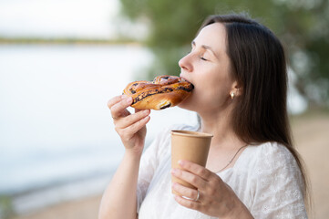 Woman enjoying sweet bun and hot drink on sandy beach, closing eyes in pleasure on summer day