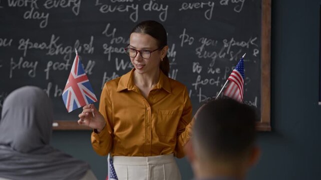 Medium shot of young Caucasian female teacher with US and British flags in hands explaining difference between two dialects to multiethnic students at their desks in modern classroom
