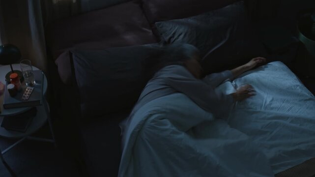 Top view time lapse of senior Caucasian woman with gray hair, in pajamas sleeping in bed at night in dark bedroom, tossing and turning, hugging pillow, covering with blanket, talking in dream