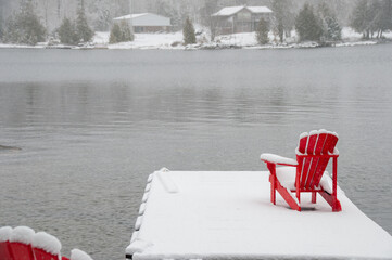Two bright red Adirondack chairs on a snow-covered floating dock on a lake. A light snowfall adds to the peaceful, serene winter atmosphere, with cottages visible on the distant shore.