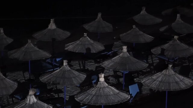 Aerial panoramic view of row of straw beach umbrellas under night lighting creating artistic shadows