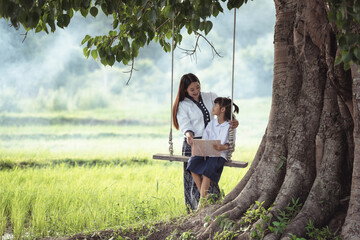 Mother and daughter enjoying a peaceful moment on a swing beneath a large tree in a serene rice field during a sunny day with lush greenery