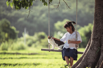 Young girl sitting on swing under tree, petting cat, in peaceful rural setting with lush green fields and soft natural light surrounding her