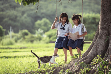 Two young girls enjoying a book while sitting on a swing under a tree in a serene rural landscape with a playful cat nearby
