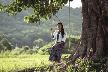 Serene Young Woman Sitting on Swing Beneath Large Tree in Lush Green Landscape with Mountains in Background on a Sunny Day