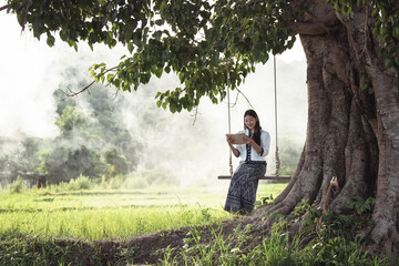 Serene Young Woman Sitting on Swing Under Large Tree in Peaceful Rural Landscape with Mist Rising in Green Rice Fields