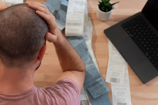 A tired man holds his head and sitting over the table with lots of bills and opened laptop while doing his tax return