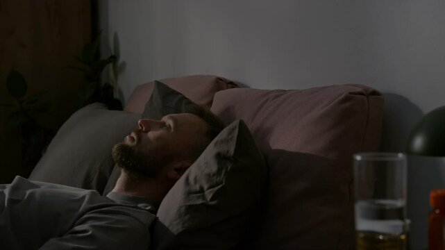 Medium time lapse of young man lying in bed at night, staring up in darkness, unable to sleep, distracted by passing car lights, sighing or doing breathing exercise