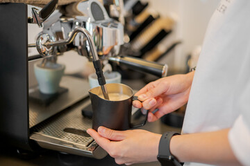 Barista steaming milk with espresso machine using black frothing pitcher. Coffee preparation process for latte or cappuccino in modern café setting