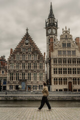 Person walking along the waterfront in front of historic guild houses and a clock tower in a European city.