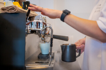 Barista using espresso machine to prepare coffee. Steam rises as milk is frothed. Ceramic cup fills below spout. Colorful cups and cloth on top. Smartwatch visible
