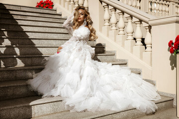 Stunning bride in luxurious white wedding gown with dramatic tulle train posing gracefully on elegant stone staircase in sunlight