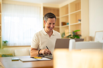 Smiling middle-aged man using graphic tablet and laptop at home office desk, working on creative project in bright modern living room.
