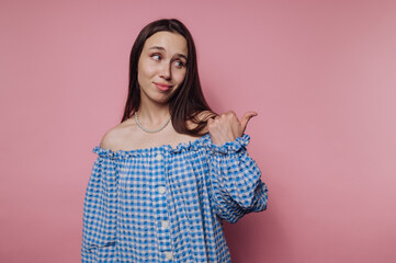 Obraz premium Young woman in blue checkered off-shoulder shirt poses against pink backdrop, casually pointing with a playful expression