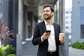 A professional man in glasses and a suit stands outdoors holding a coffee and smartphone, smiling confidently. The background features a sleek, modern architectural design and blurred greenery.