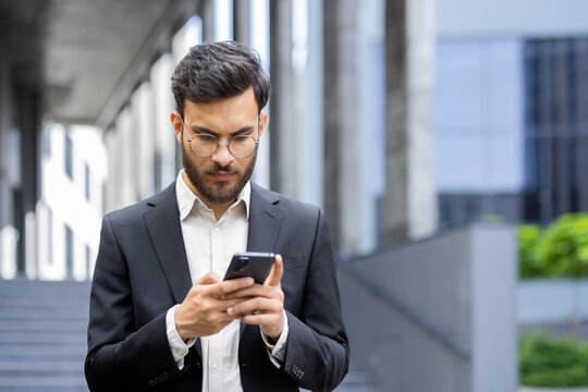 A professionally dressed man engrossed in using his smartphone while standing outside an office.