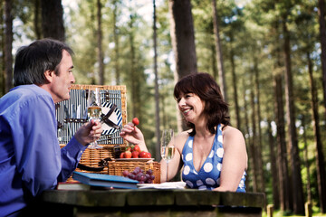 A mature couple enjoying a picnic in a forest park. They are seated at a wooden table, holding wine glasses, and smiling at each other with a large wicker basket between them. From a series of images.
