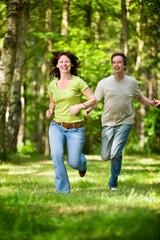 Fototapeta premium A happy mature couple run together through a sun-dappled forest path smiling and enjoying their time outdoors together. Part of a series of images.