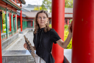Elegant woman in black dress with decorative fan posing in colorful Chinese-style pavilion with red columns and green landscape in background