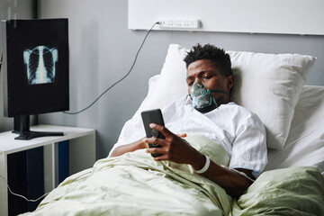 Horizontal shot of young African American man with IMV mask on face lying in hospital bed surfing Internet on smartphone