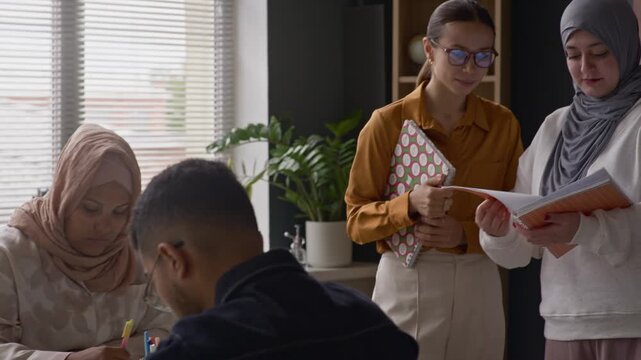 Muslim student in hijab showing copybook to Caucasian female teacher while diverse classmates writing at table during English lesson in modern, multicultural classroom