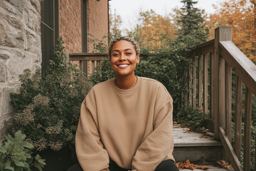 Smiling woman in plain tan crewneck sweatshirt sitting outdoors on wooden porch steps with autumn foliage in background | Lifestyle portrait | Apparel catalog or branding use