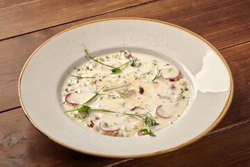 Cold okroshka soup with vegetables and herbs served in white plate on wooden table
