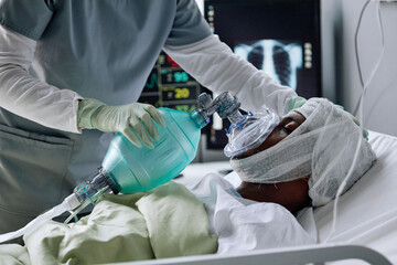 Unrecognizable nurse saving life of injured young Black man setting mechanical ventilation for him in hospital ward