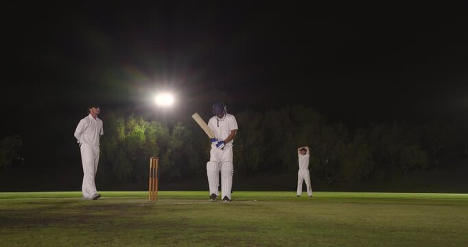 Glancing at floodlight diverse male cricketers bowling ball swinging bat catching and returning it