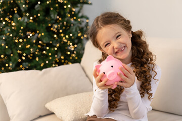 Banking, finances, investment and happy people concept. Cute happy little girl sits on sofa and holds pink piggy bank in her hands on Christmas. Pocket money, family budget and savings concept.