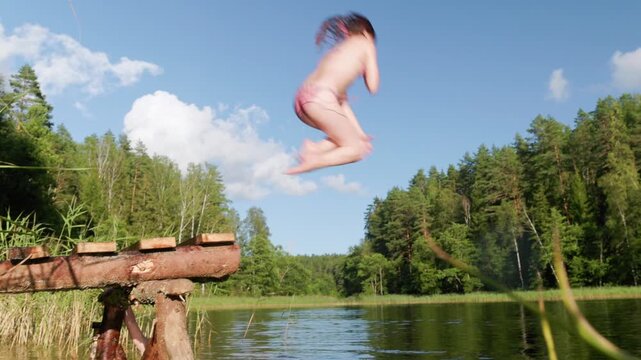 Young girl in a swimsuit leaps from a rustic wooden pier into a forest lake, creating a big splash on a bright summer afternoon surrounded by nature