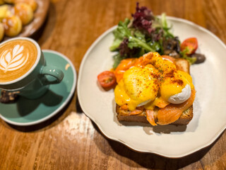 Brunch plate with smoked salmon eggs benedict topped with hollandaise sauce, served with salad and cherry tomatoes, alongside a cappuccino with latte art on a wooden cafe table.