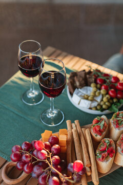 Overhead view of tomato bruschetta, breadsticks, grapes, cherry tomatoes, Camembert cheese, brown bread, olives and two glasses of red wine on a garden table