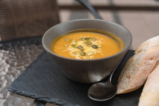 Close-up of a bowl of pumpkin soup with pumpkins seeds, grated cheese and slices of fresh ciabatta bread on a garden table