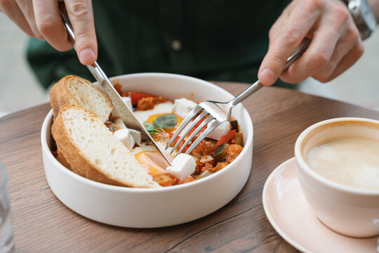 Close-up of a man sitting at a table eating shakshouka with poached eggs, feta cheese, sliced bread and a latte coffee