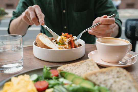 Close-up of a man sitting at a table eating shakshouka with eggs, sliced bread, a glass of water and latte coffee
