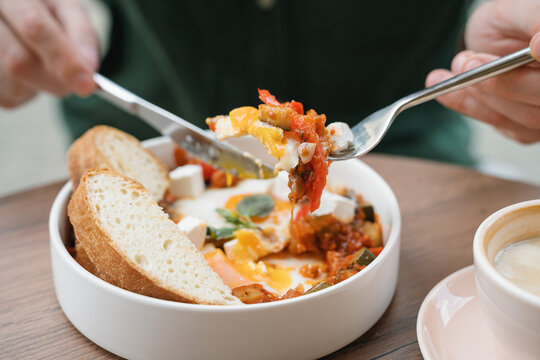 Close-up of a man sitting at a table eating shakshouka with eggs, sliced bread and a latte coffee