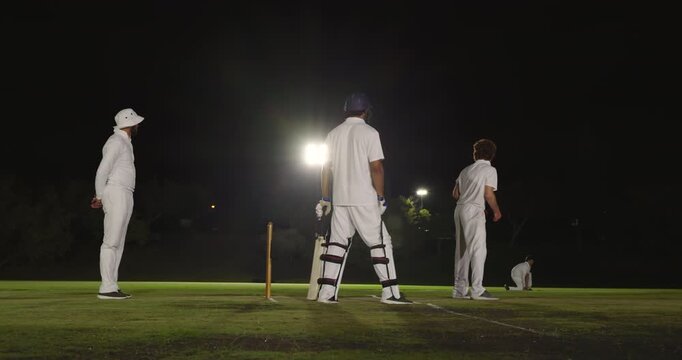 Initiating delivery, male bowler running under floodlights bowling at stumps while umpire observing