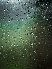 Rain-soaked window with droplets streaming down the glass. Beyond the window, the view is blurred, revealing indistinct foliage, indicating a rainy day. The overall mood is calm and reflective. High