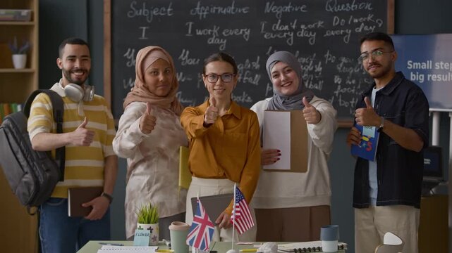 Medium portrait of smiling teacher and diverse students posing in front of chalkboard, showing thumbs up during English lesson in multicultural classroom