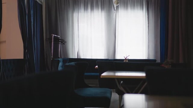 Interior of modern caf with velvet blue seating and a small table by large window. Curtains and minimalist d cor create a calm, peaceful atmosphere. Light filtering through the window