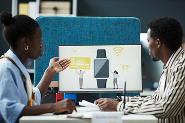 Black woman and man discussing digital banking solutions in bank office, gesturing toward computer monitor displaying smartwatch with credit card and online payment icons