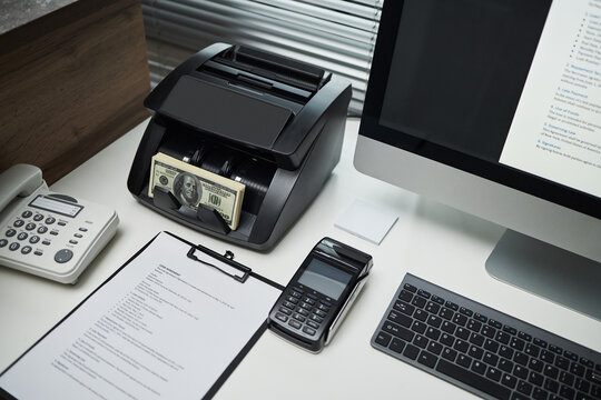 Office desk showing money counting machine processing US dollar bills beside desktop computer, calculator, landline phone and clipboard, illustrating bank office financial operations - Powered by Adobe