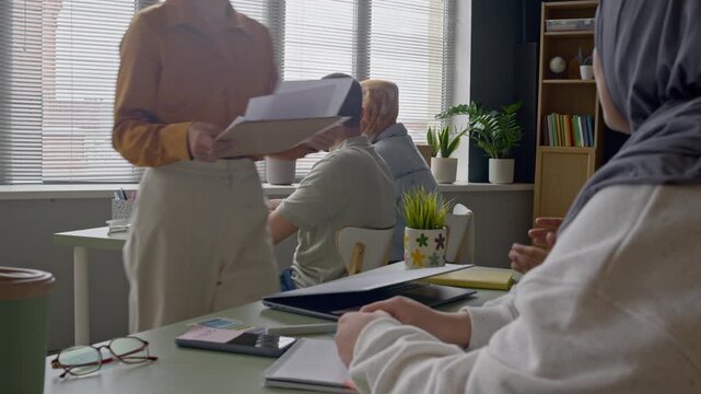 Teacher handing out worksheets to diverse students seated at desks during English lesson in modern classroom