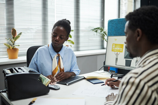 Black woman counting cash at desk while African American male client sitting across reviewing documents in bank office, computer monitor displaying credit card image, indoor business setting - Powered by Adobe