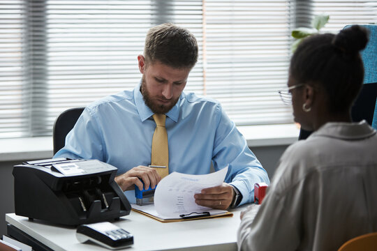 Caucasian male consultant reviewing documents with Black young woman sitting across desk in bank office, money counting machine and paperwork visible on table, both focused on discussion