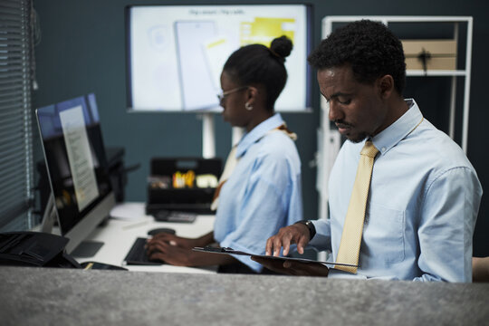 Black male bank employee reviewing documents on clipboard while Black woman working on computer in modern bank office with digital screens in background